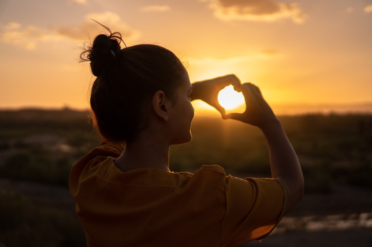 Woman watching sunset making heart shape with her fingers.