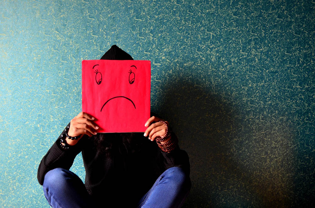 Man holds up a red card in front of his face which has a sad face drawn upon it